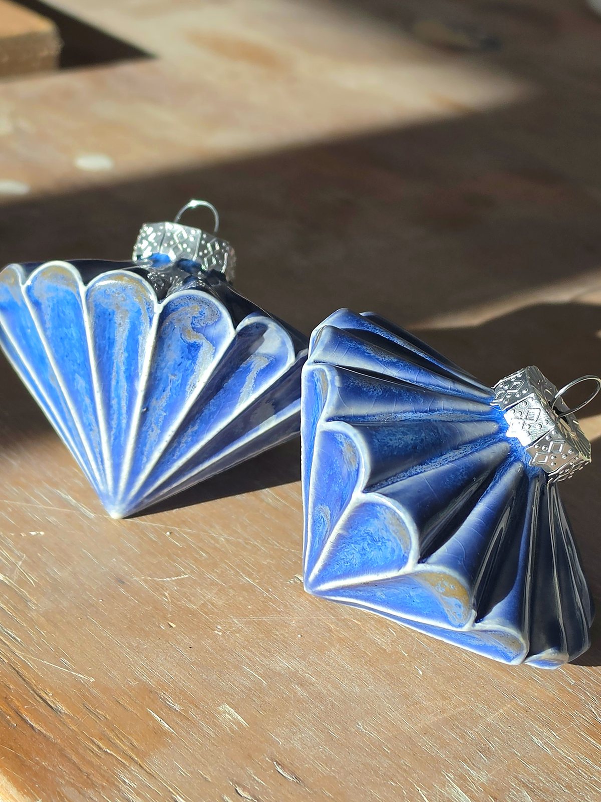 Two Floating Blue ceramic baubles resting on a sunlit workbench, showing the pleated cone form and silver-tone caps.