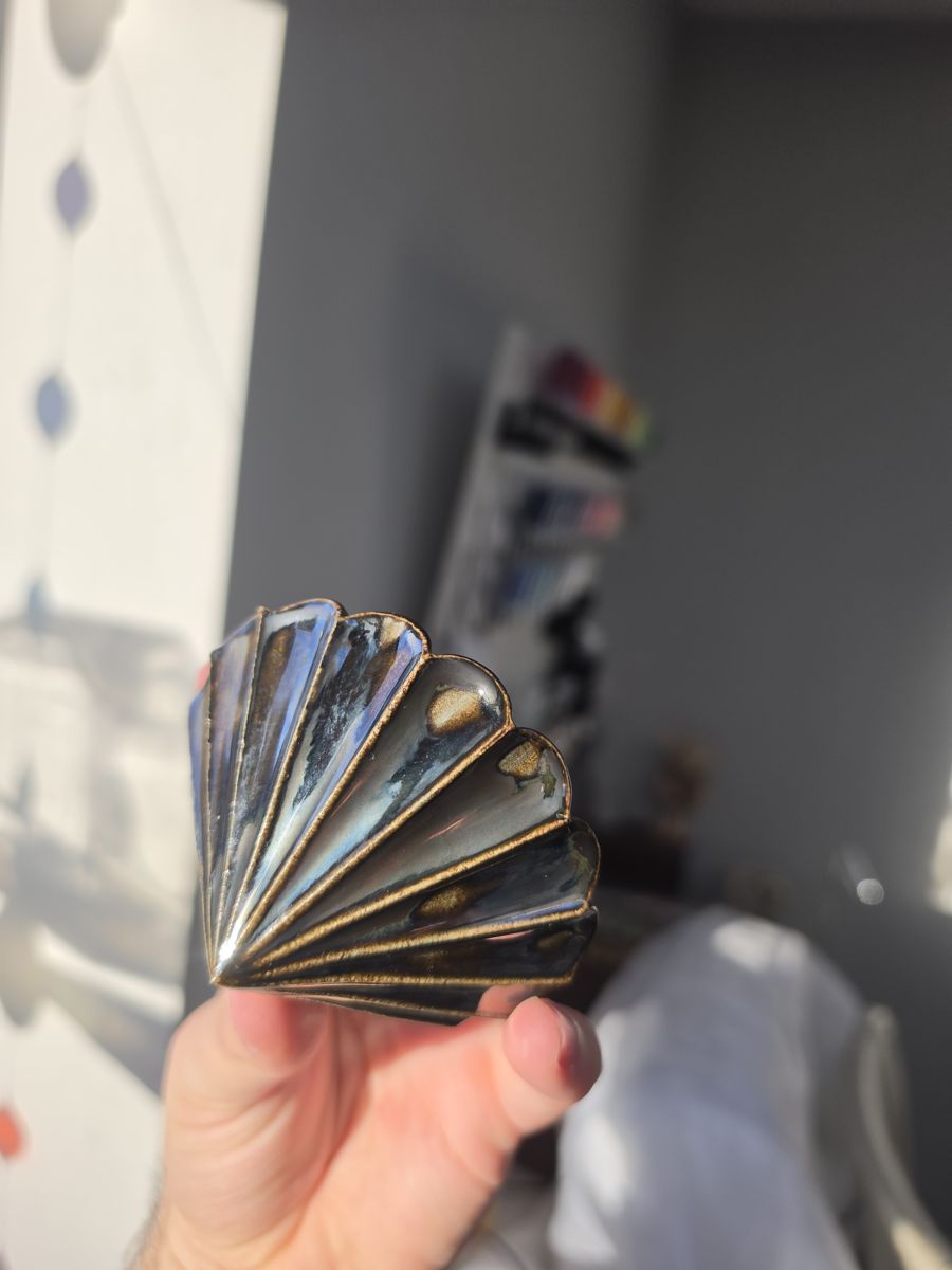 Black metallic fan-shaped ceramic bauble held in a hand, dark glaze with bronze edges on each pleat.